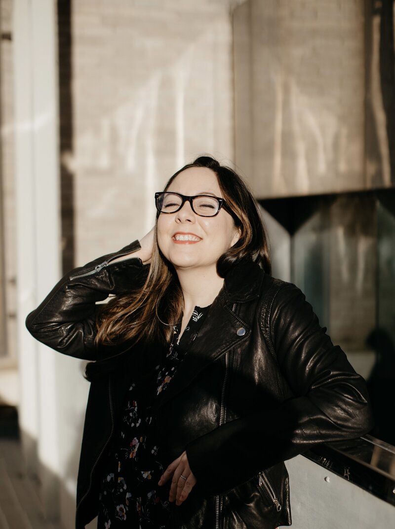 A woman with brown hair and glasses wears a black leather jacket and smiles at the camera gleefully.