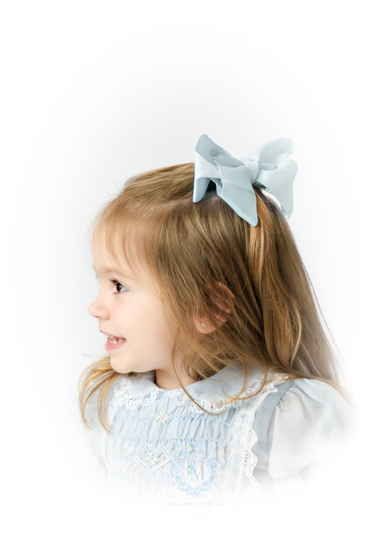Toddler girl with long brown hair, side portrait in a vintage blue dress with a lace collar and blue bow.