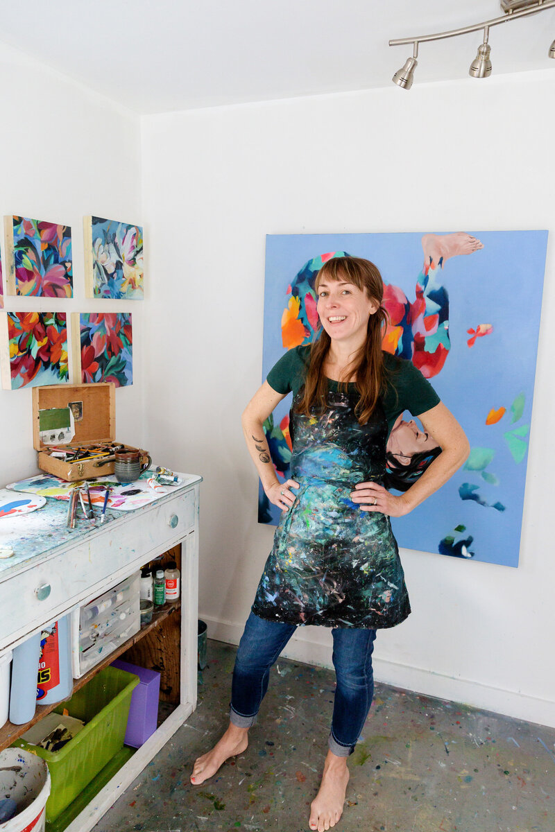 Calgary brand storytelling photoshoot of a barefoot female artist standing confidently in her studio, wearing a paint-splattered apron over jeans and a dark green top. She is surrounded by her vibrant, abstract artwork, with a large canvas featuring a colourful, surreal figure behind her. A well-used workbench holds paint tubes, brushes, and an open wooden box filled with art supplies, showcasing her creative process.
