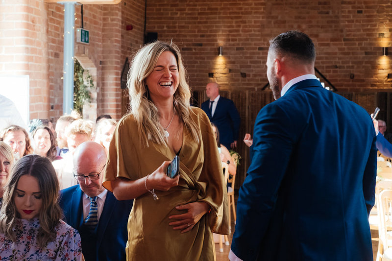 blonde haired lady dressed ina gold dress, stands in wedding ceremoney room and laughts hard whilst holding her stomach at Swallows nest wedding barn