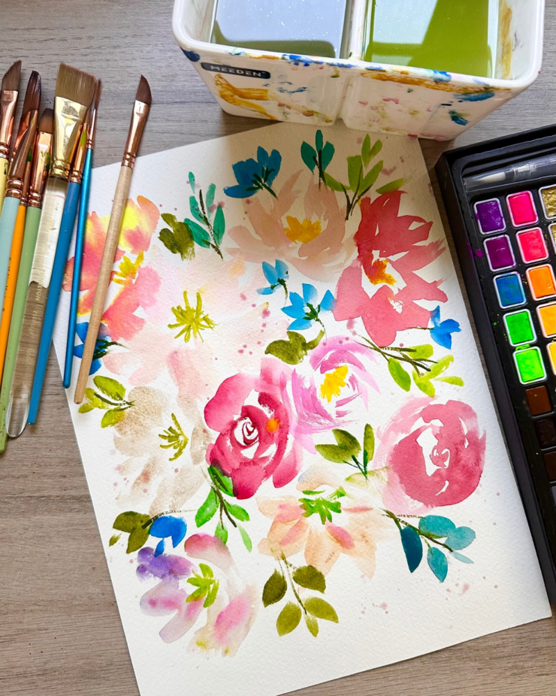 Colorful watercolor flower painting surrounded by paintbrushes and a paint palette