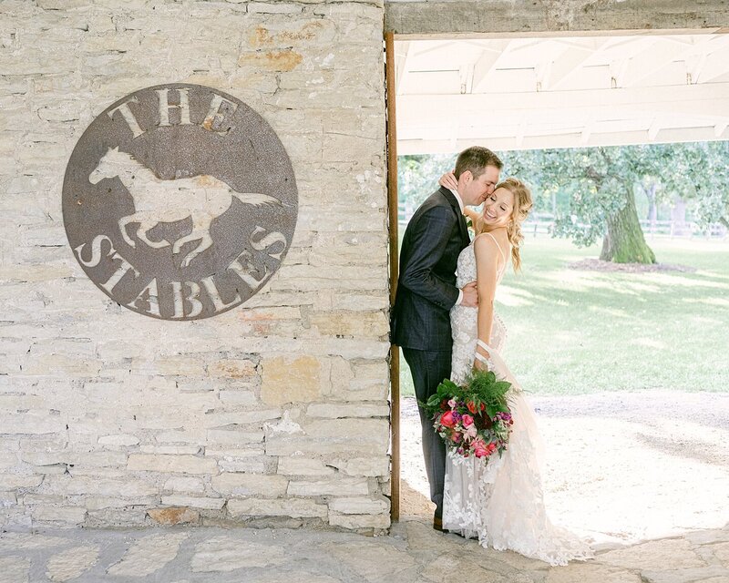A groom kisses his smiling bride on the cheek as she looks down at the ground. They are in the old stables on the grounds of Mayowood Stone Barn in Rochester, Minnesota, following their wedding.