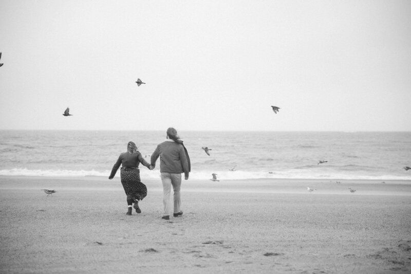  A couple walks hand in hand on the beach, with birds flying overhead, capturing a serene moment by the water.
