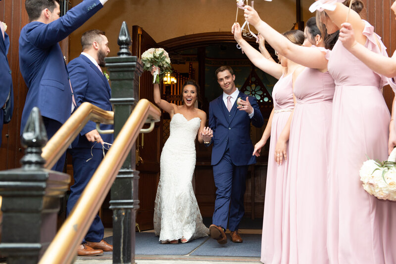 Excited and happy bride and groom exit a church in Cleveland Ohio, holding hands as they celebrate their wedding surrounded by family, friends and loved ones cheering them on. Photographed by Aaron Aldhizer