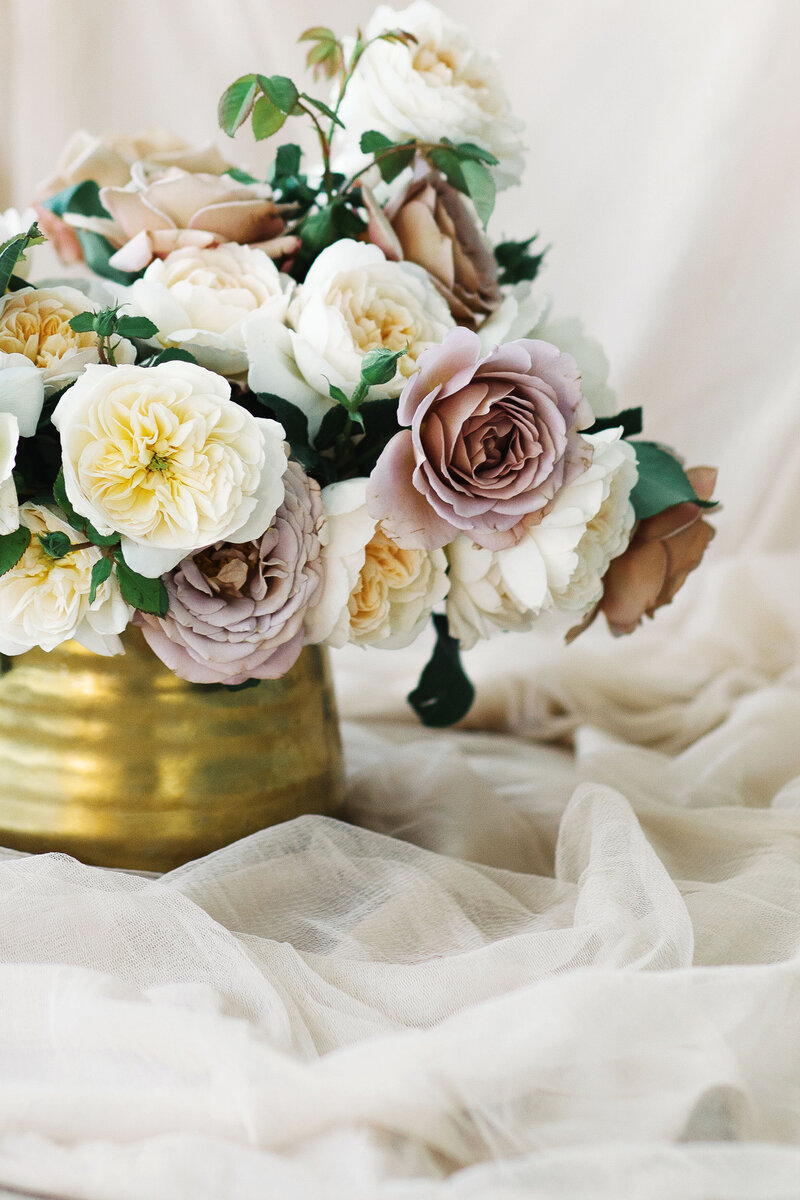 Gold vase with pale pink roses and white peonies sitting on beige coloured sheer fabric