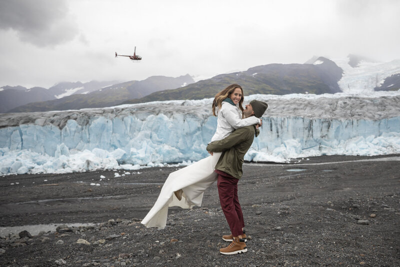 Groom holds is new bride in the air while a helicopter flys overhead. They stand in front of a huge glacier after the heli dropped them off here for a ceremony. 