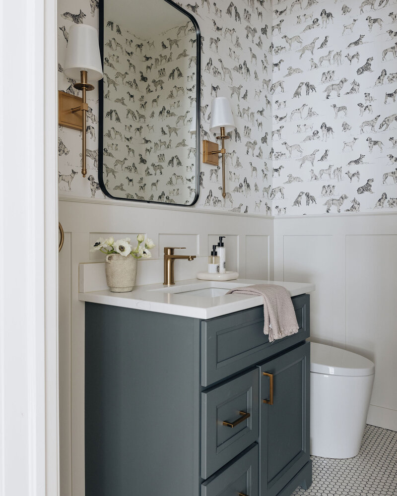 Stylish powder room in Montreal featuring playful wallpaper, gold fixtures, navy vanity, and a curved black mirror – by Ingrid Interior Design.