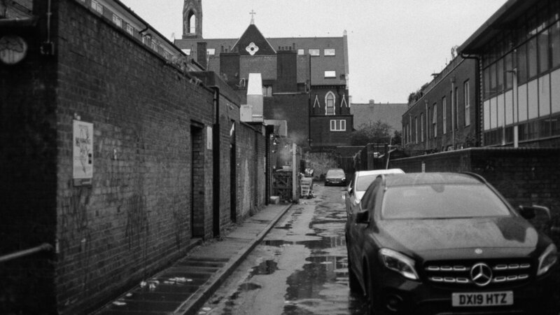 Black-and-white photo of a narrow, wet alley lined with brick buildings, a parked Mercedes in the foreground, and an old Gothic-style building with a cross in the background.