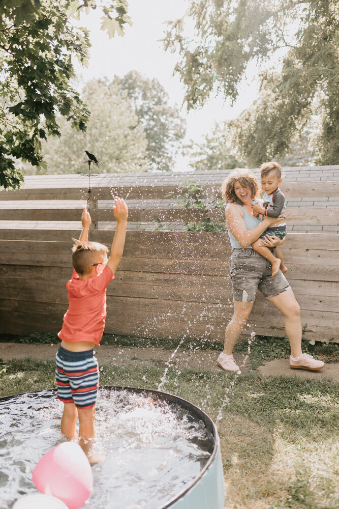 A family photographer in West Lafayette captures a small boy in a backyard pool splashing his mother while she joyfully runs past. 
