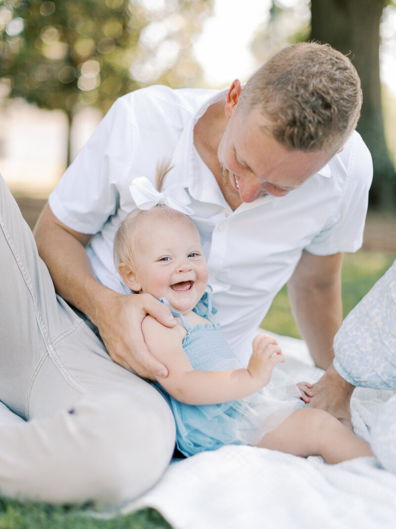 Blonde dad in white shirt holds one year old daughter in blue dress as they laugh together, taken by Little Rock photographer Bailey Feeler