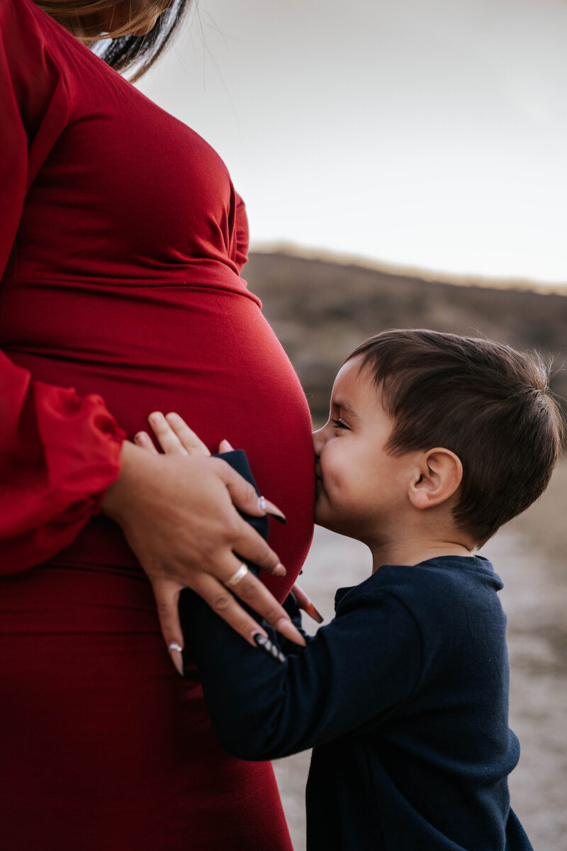A side profile  maternity photo in a dry field of a mother in a red dress and her four year old son kissing his mother's baby bump that holds his soon-to-be baby sister