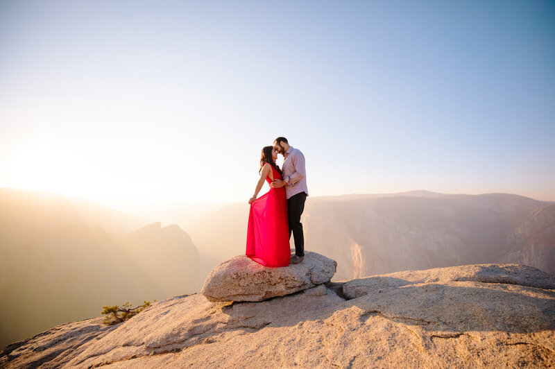 Man and woman embrace as woman holds her dress as they stand on a rock  surface during sunset