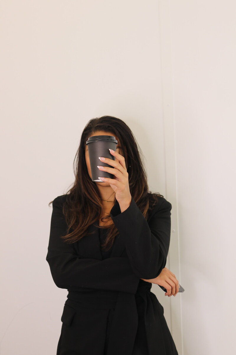 Woman in a black outfit holds a black coffee cup covering her face, standing against a plain white background, conveying mystery and anonymity.