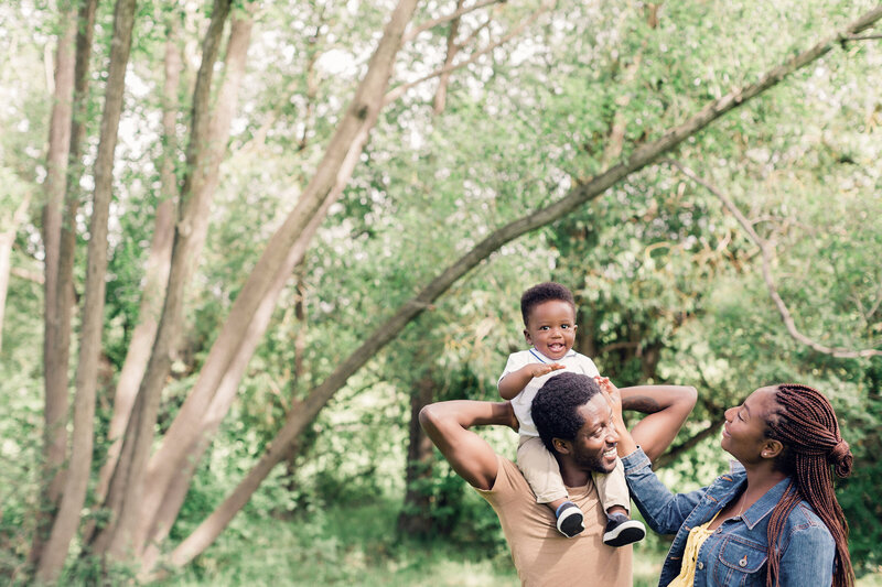 A joyful toddler sits on his father’s shoulders while the family stands together in a lush park – Family Autism Support Coach at Chantal Hewitt Coaching