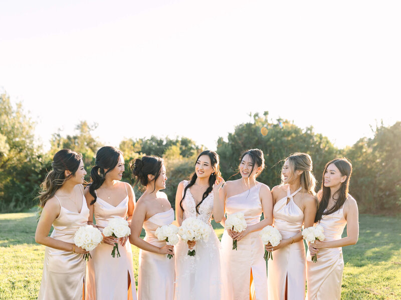 Bride in white gown and bridesmaids in pale pink dresses stand on a sunny grassy field, holding white bouquets, smiling and laughing together.
