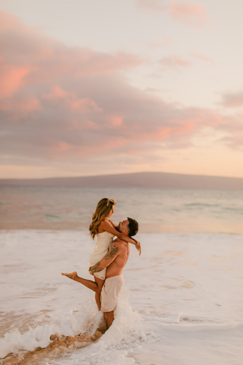 sunset engagement portrait featuring man and woman cuddling as tide washes over their feet