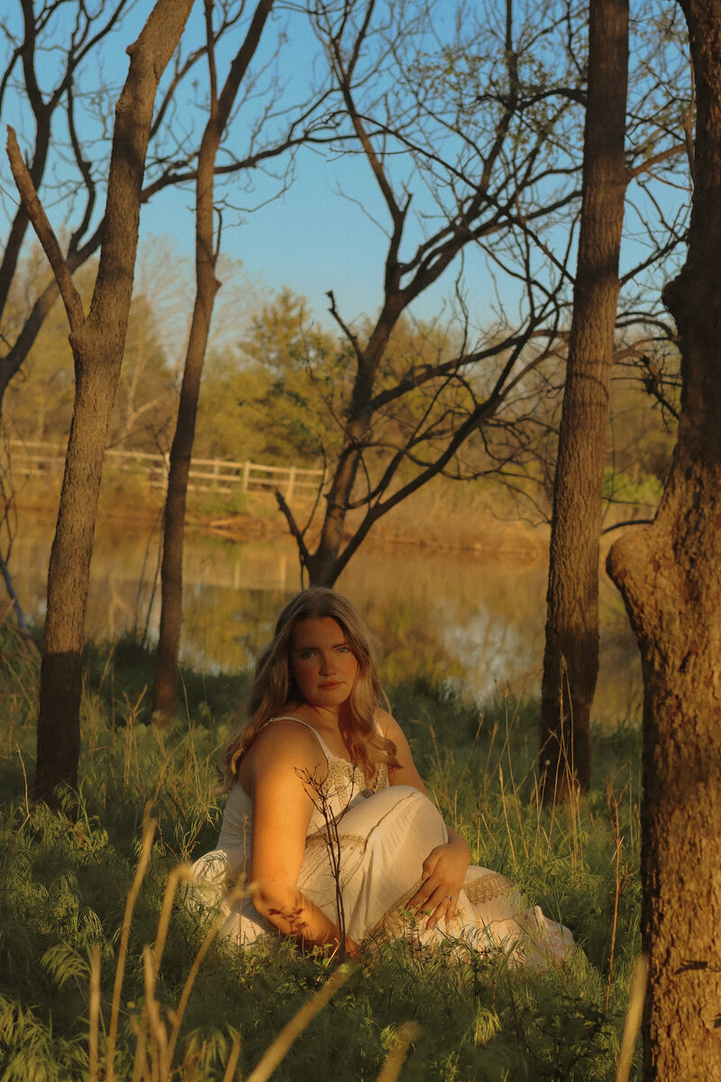 A woman is sitting in a field of tall grass and flowers while the sun illuminates her through the trees. A pond can be seen in the background 