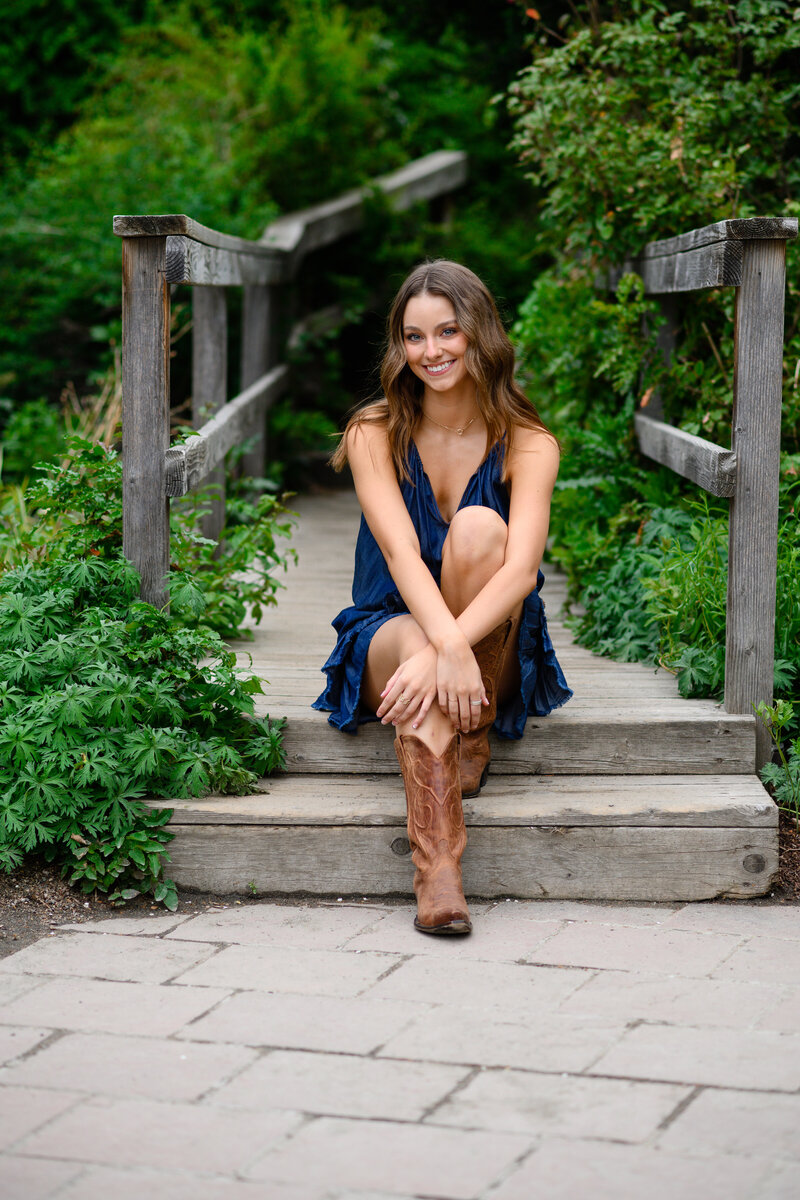 A denver senior girl with cowboy boots on sits on a wooden bridge at the denver botanic gardens posing for Denver senior photographer