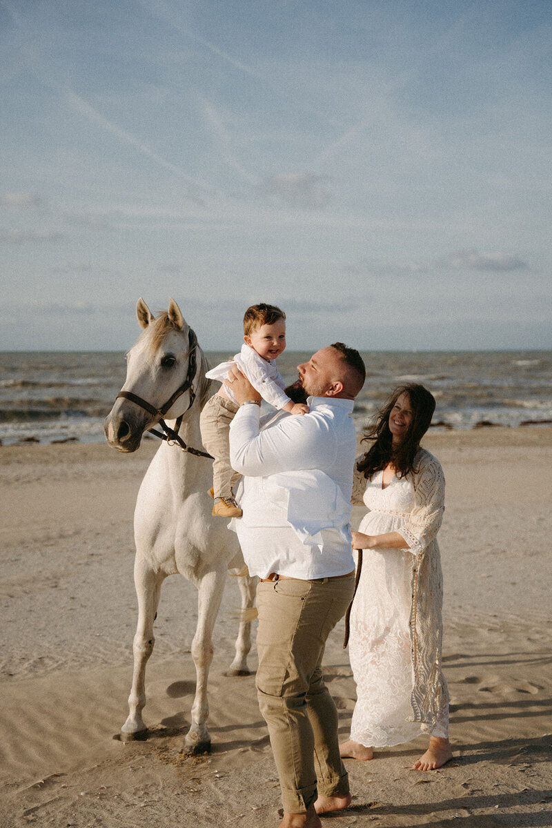 Famille posant à la plage avec leur petit garçon et leur cheval blanc. Séance photo famille et grossesse.