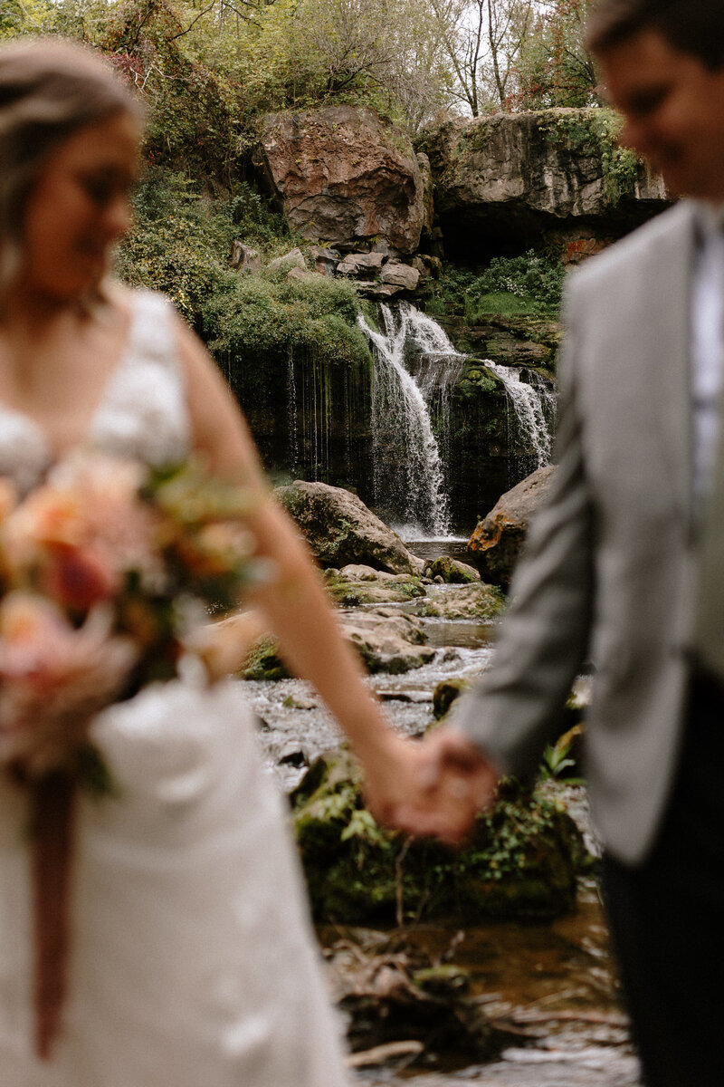 A couple holds hands in front of a picturesque waterfall surrounded by lush greenery and rocky terrain, capturing a romantic moment.