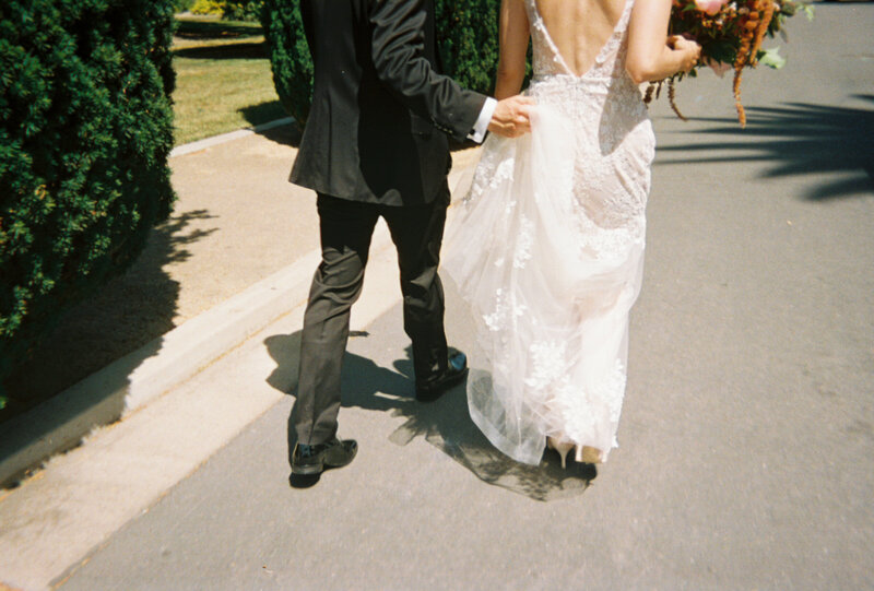 Film photo of bride and groom walking together while groom holds up bride's dress