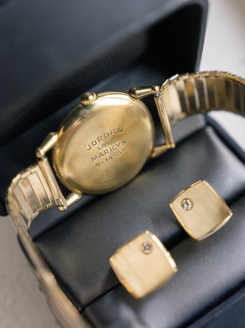 A gold heirloom wristwatch with an inscription reading Jordan Loves Marilyn 12-25-57 is displayed in an open black box alongside a pair of square gold cufflinks with small embedded gems.