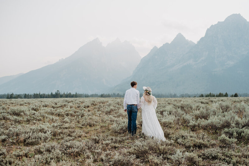 jackson hole wedding with bride and groom holding hands and walking in a sage brush field in the tetons with a light fog over the mountains