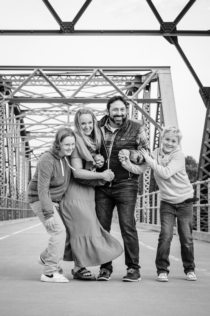 A joyful family of four poses on a metal bridge, with everyone leaning into each other and laughing, captured in black and white for a timeless feel.