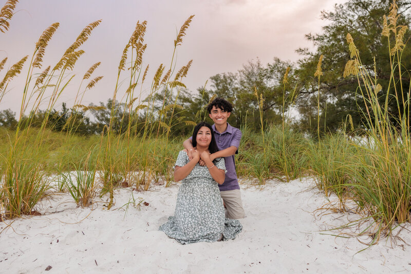 Mother and son kneeling on the sand with beach grass behind them. the son has his arms wrapped around moms neck.
