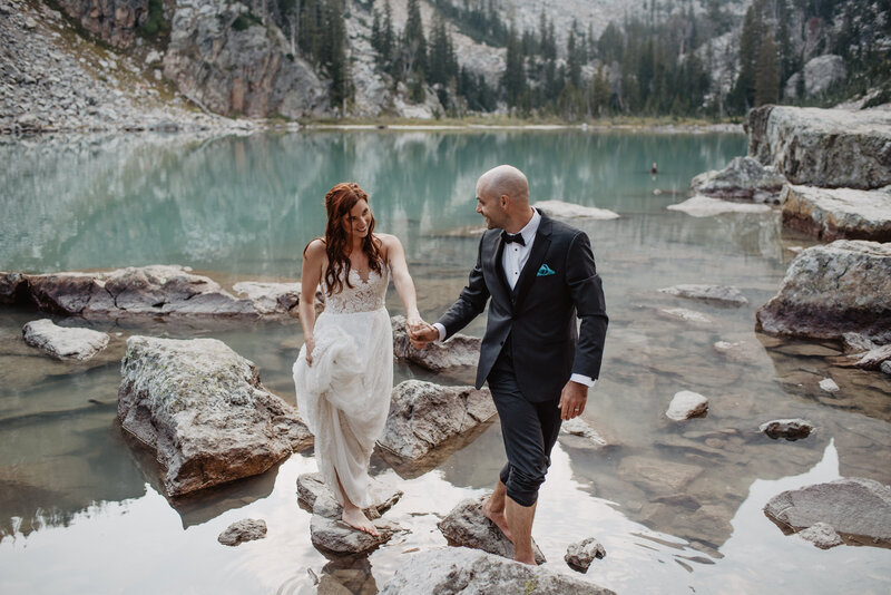 jackson hole photographers photographs bride and groom holding hands and walking from one rock to another at Delta Lake