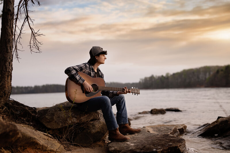 Atlanta Senior photo of boy sitting on rock by the lake, playing a guitar. Photo was taken by Jenni Nikki Photography who does Senior and graduation photography.