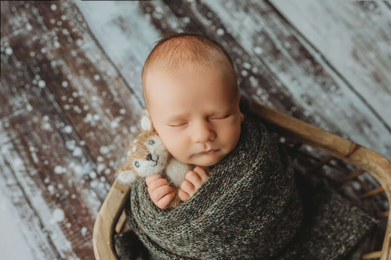 A serene newborn baby is sleeping, wrapped snugly in a soft, neutral-toned fabric. The baby has closed eyes and a calm expression, lying against a gentle, textured backdrop that enhances the cozy atmosphere.