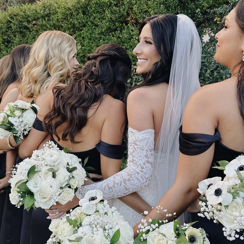 Bride in white gown with sleeves holding bouquet among her bridesmaids dressed in satin black dresses in the gardens of La Valencia Hotel.