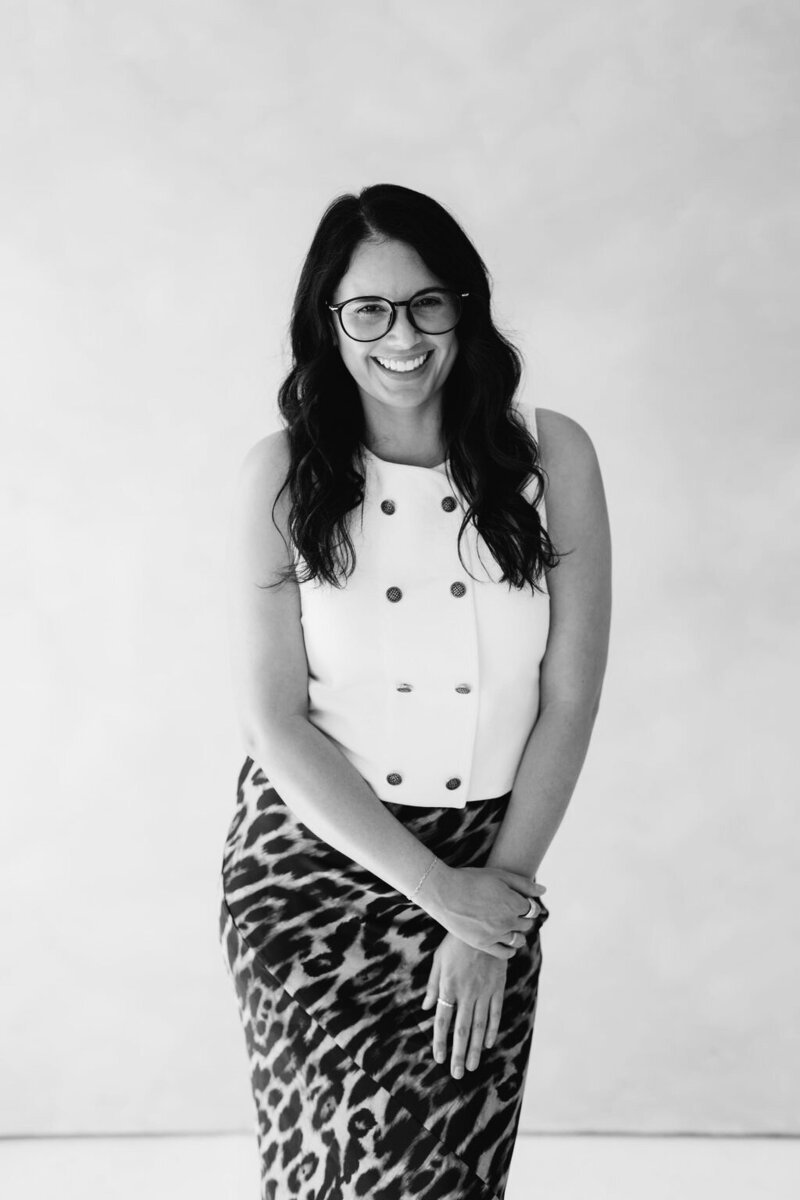 Raquel Bickford, founder of La Perle Retreats and ROQUE Events, smiles confidently in a black-and-white portrait, surrounded by elegant florals. Photo by Logan Alexander.