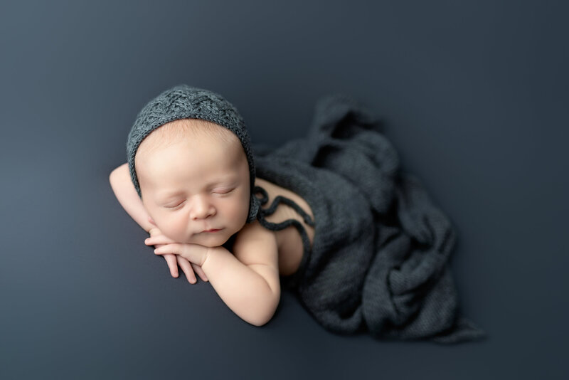 Classic newborn pose with textured knit bonnet on rich grey – softly lit and timeless.
