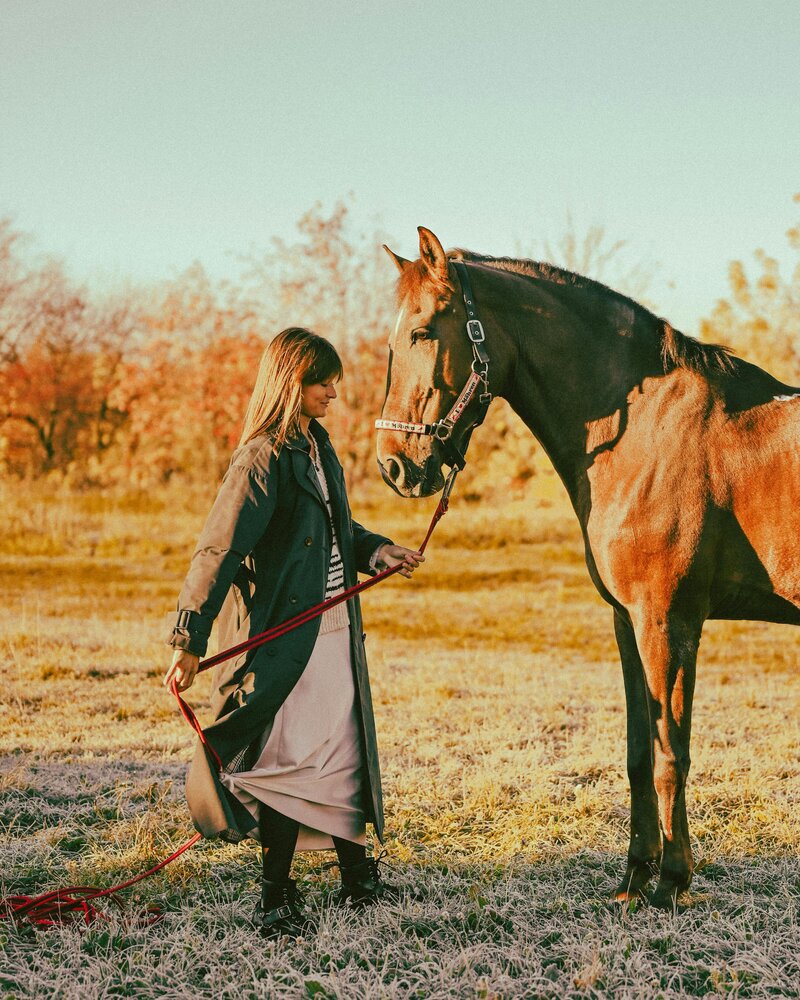A woman wearing a long dress and coat faces a horse in a field, holding into a lead