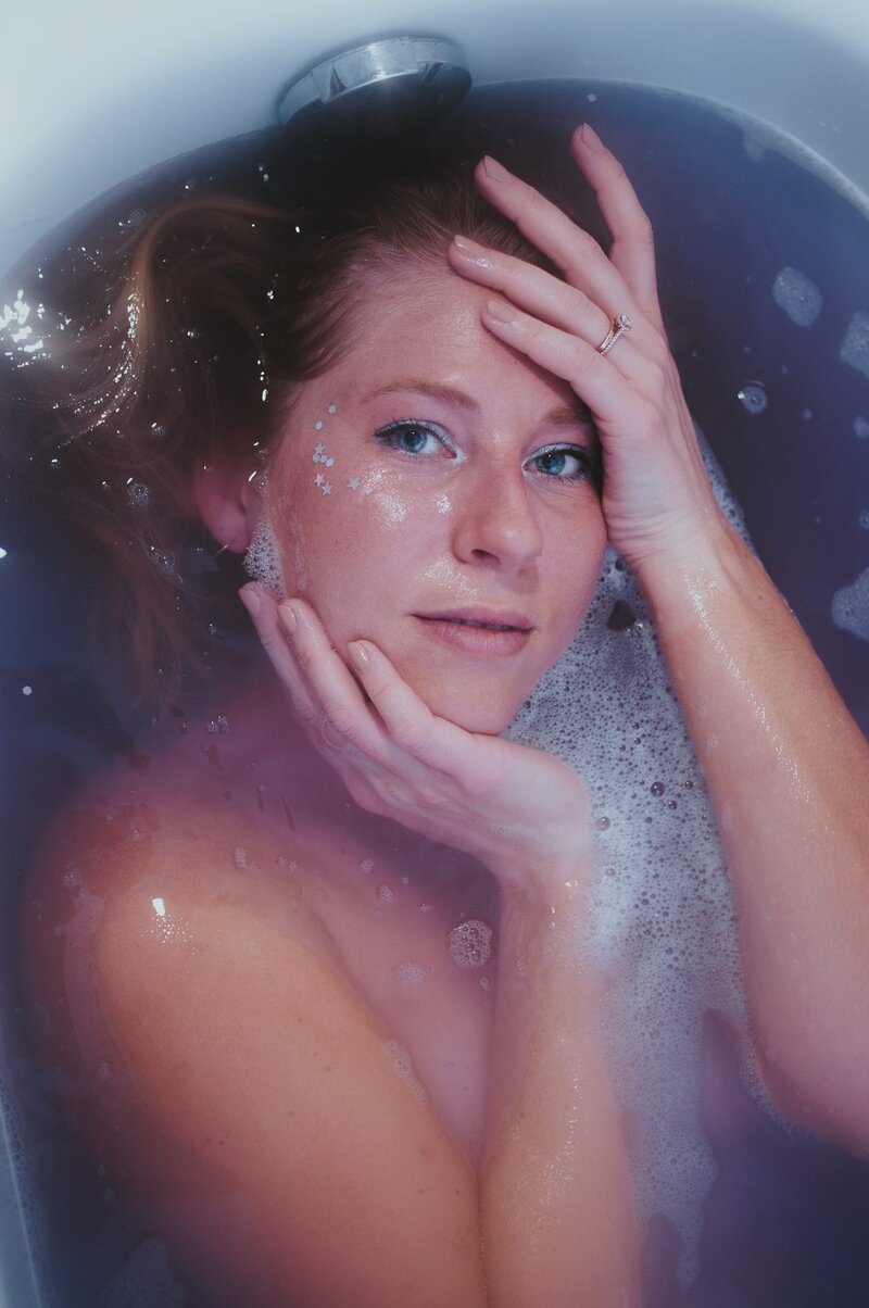 Woman with sparkles on her face poses in sudsy bathtub water.