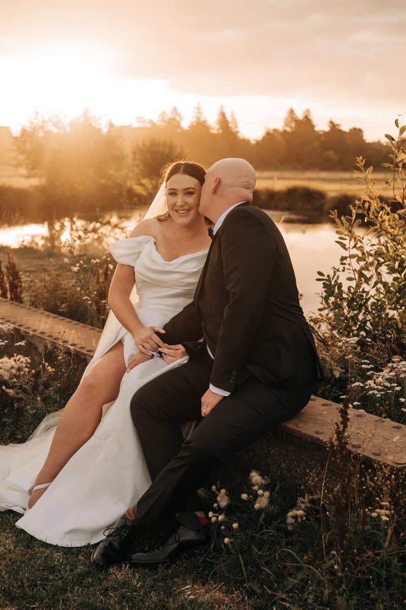 newly married couple sit on bench beside pond at sunset in paperswan dress in darfield at bangor farm