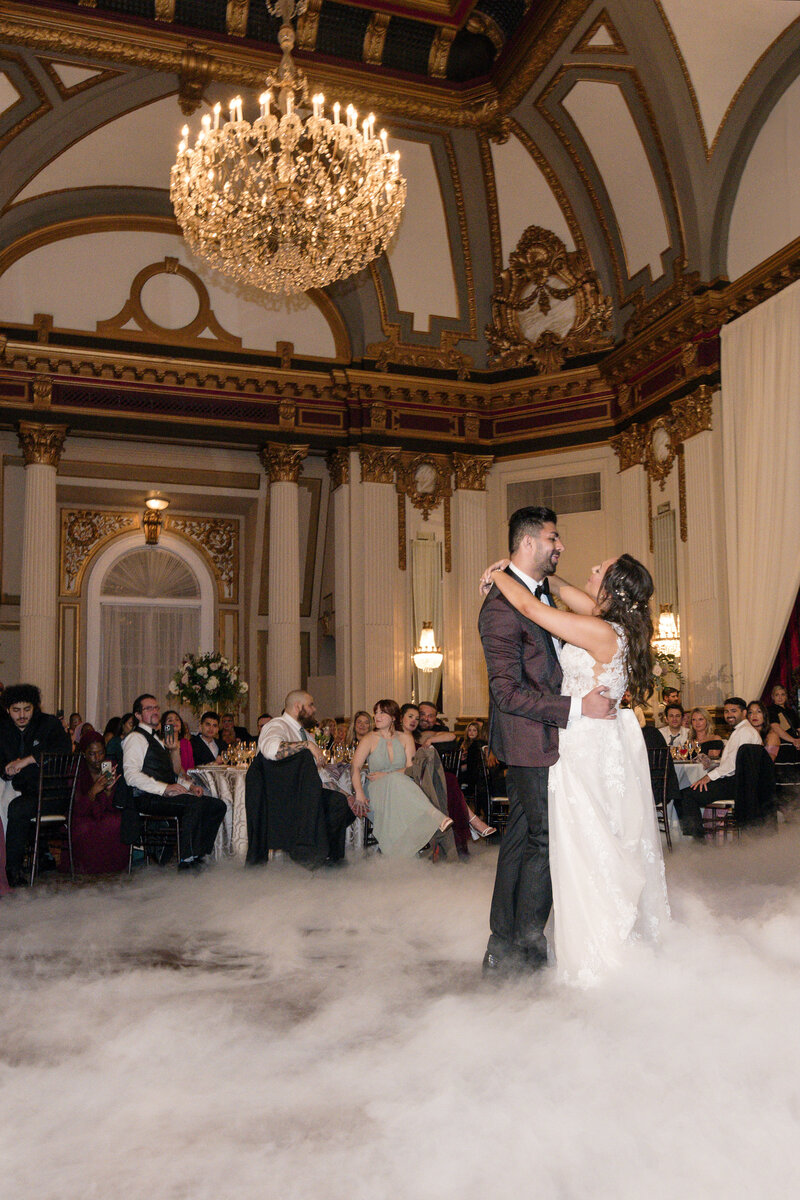 A couple shares their first dance at a wedding reception in an opulent ballroom with a grand chandelier and ornate decorations. Guests sit around tables in the background, and a misty fog covers the dance floor.