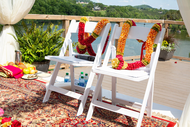 Two white chairs decorated with colorful floral garlands on a deck overlooking a river in Iowa, set up for a stress-free wedding event.