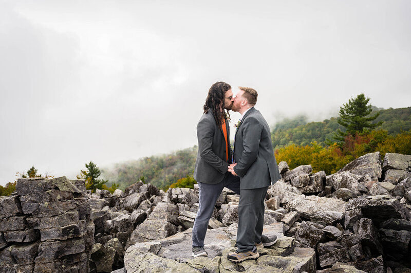 A queer couple stands on a pile of rocks at Blackrock Summit during their elopement day in Shenandoah National Park. The pair hold onto one another's hands and go in for their first kiss at the conclusion of their fall wedding ceremony.
