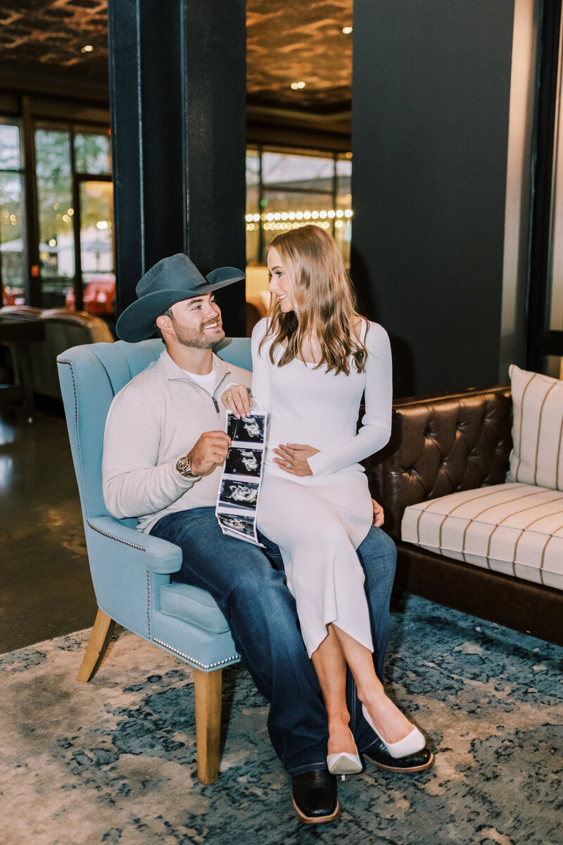 Woman sitting in her husband's lap in a blue chair while they hold up their pregnancy ultrasound pictures