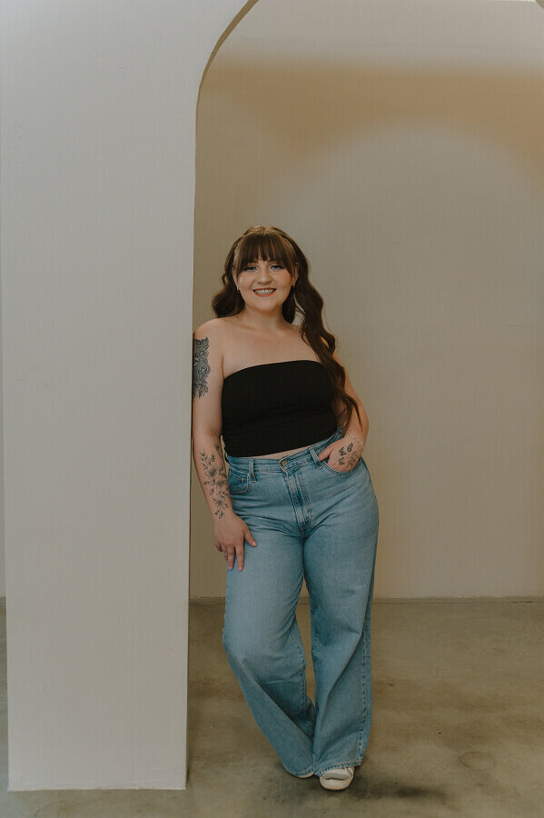 Leaning against a beige wall, a young woman smiles for a business branding session. She's wearing a black tube top and blue jeans.