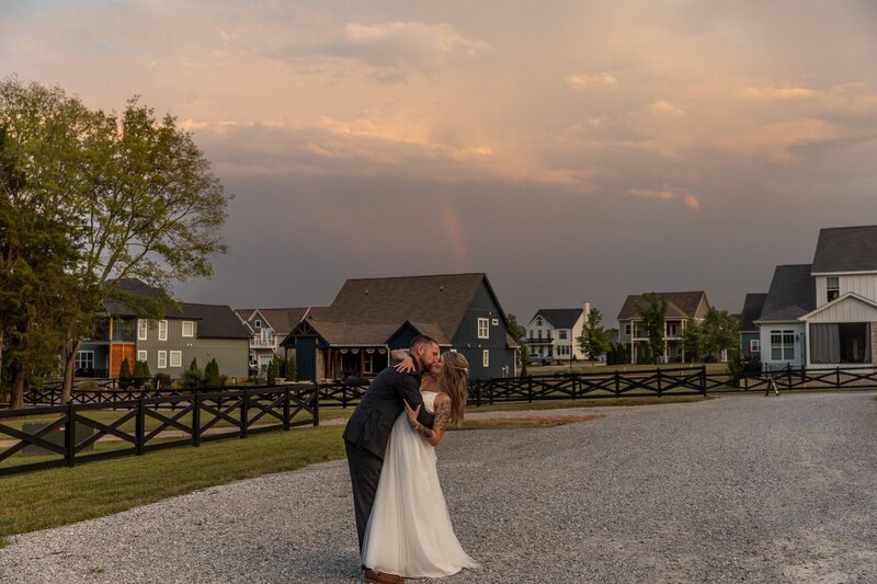 Bride and groom embrace and kiss passionately under moody skies and a rainbow, there is a black fence on the left of the frame and houses in the background