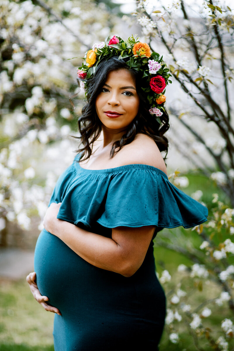 a pregnant woman in a teal dress and flower crown stands in front of a flowering tree in boulder, colorado