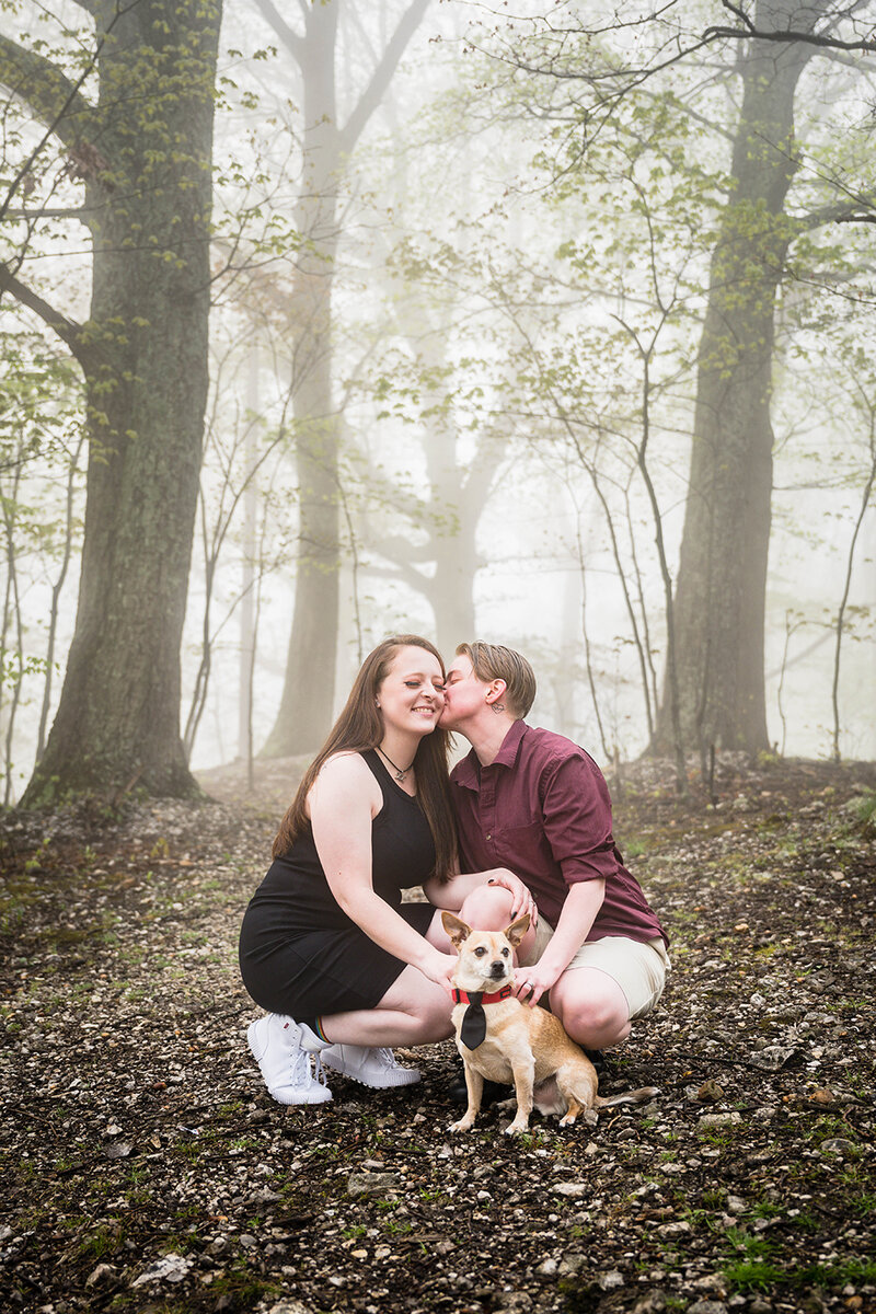 A couple squats with their dog during their engagement portrait adventure session at Mill Mountain Park in Roanoke, Virginia. A partner kisses the cheek of their significant other while the smile widely with their eyes closed.