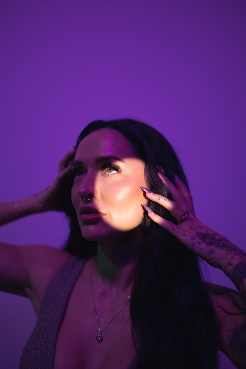 Woman with dark, long hair and tattoos on her arm looks off-camera with a spotlight on her face and moody purple background. She holds her hands up to her face to pose with long purple nails.