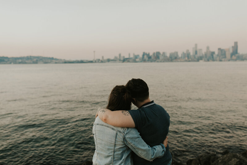 A couple sits with their backs to the camera, arms around each other, looking out at the water and city skyline.