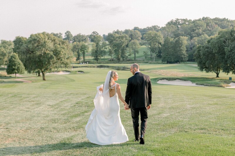 Bride and Groom walking with back towards camera. They are in a golf course field on a sunny day.