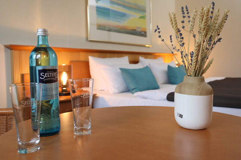 Tisch mit Vase, einem Glas sowie einer inkludierten Flasche Wasser und das Bett im Zimmer.